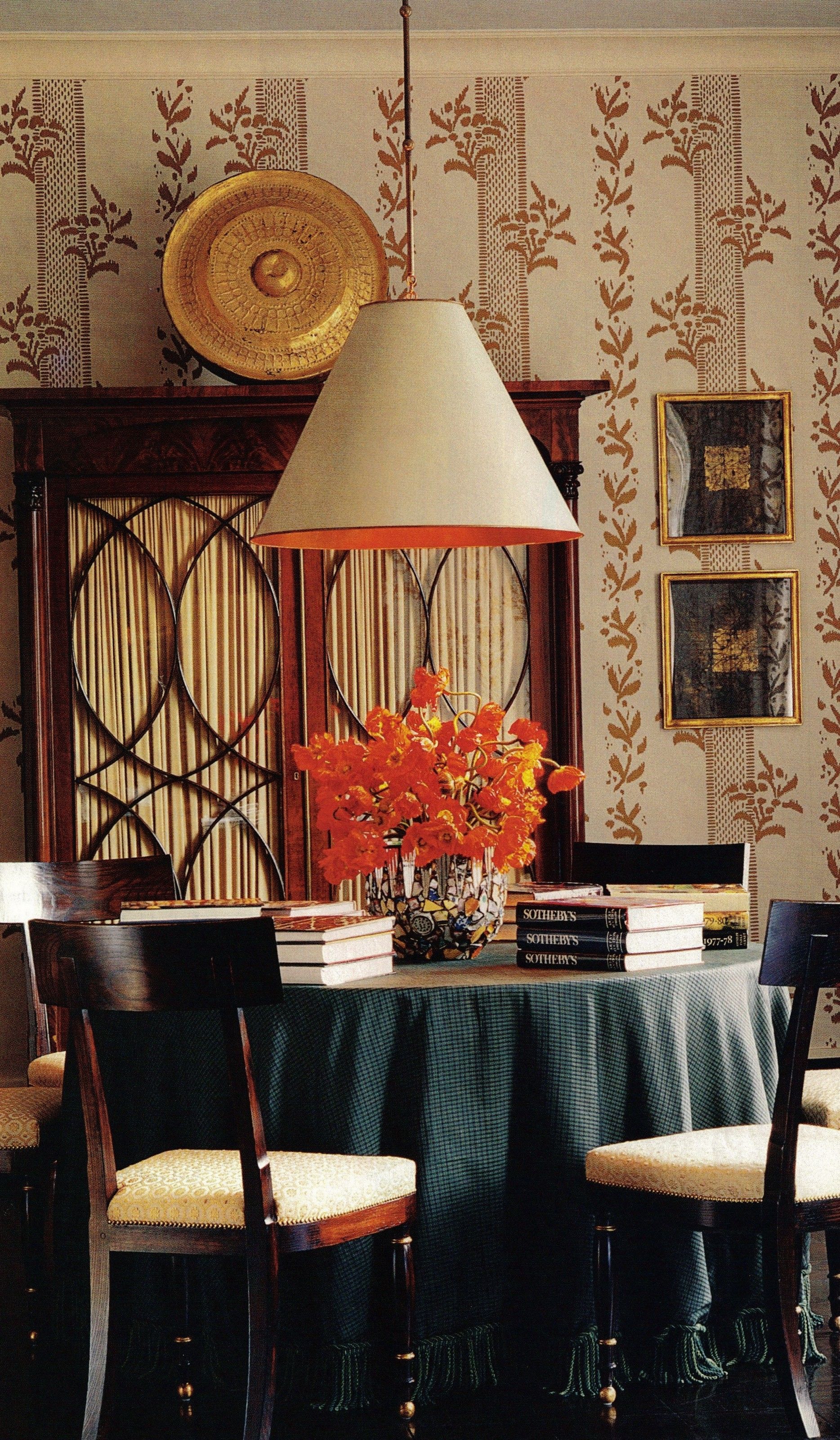 A dining room by Albert Hadley. It has a light blue ceiling, a favourite Hadley hue for the upper plane. The American Empire mahogany armoire is topped by a Tibetan gong. Next to them are two works on paper by Connecticut artist Mark Sciarillo, also a metalworker, who made the sculpted bronze base of the living room's coffee table. The vellum lampshade, the Eyelet gold-on-ivory wallpaper, and the chairs are all Hadley's designs. Fernando Bengoechega photography. House Beautiful